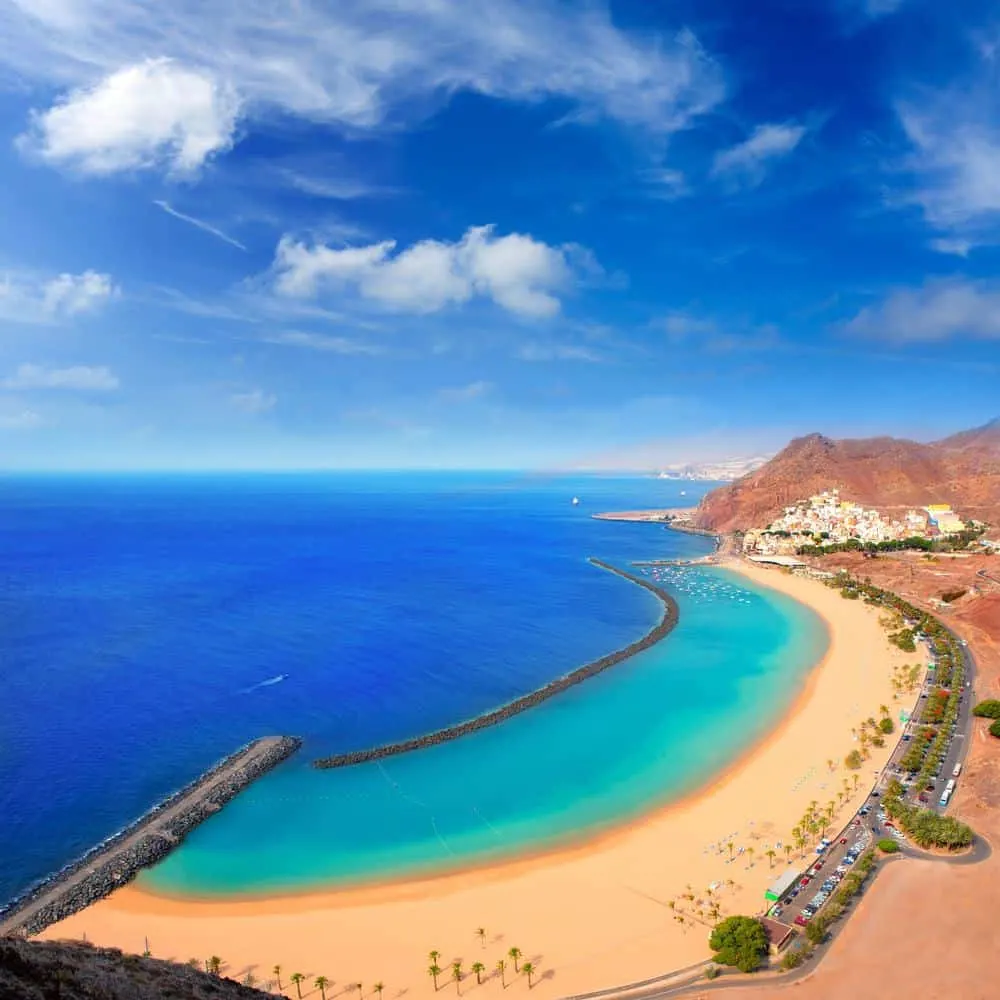 best beaches in tenerife south, vView down towards large golden yellow sandy beach with turquoise and rich blue sea water on one side and palm trees and parked cars on the other with a built up area nestled into the crook of a tall rocky hill in the distance all under a blue sky with white wispy clouds