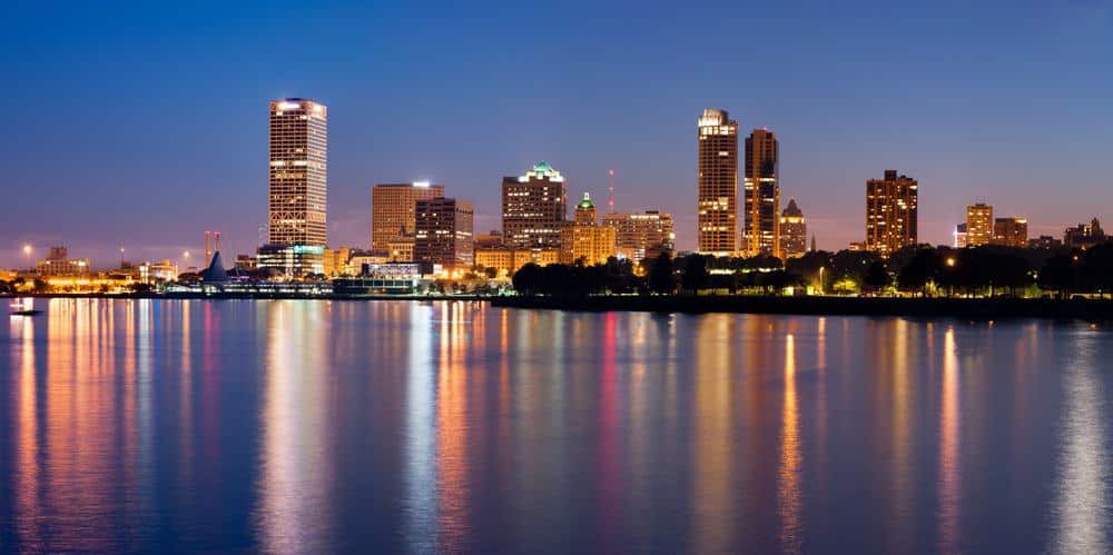 skyline view of Milwaukee at sunset with a series of tall skyscrapers lit by sporadic lights standing on the banks of a large body of water under a clear sky