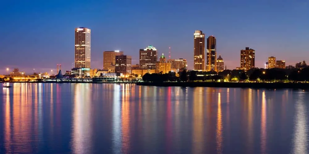 Get involved in some Wisconsin Fall Getaways, skyline view of Milwaukee at sunset with a series of tall skyscrapers lit by sporadic lights standing on the banks of a large body of water under a clear sky