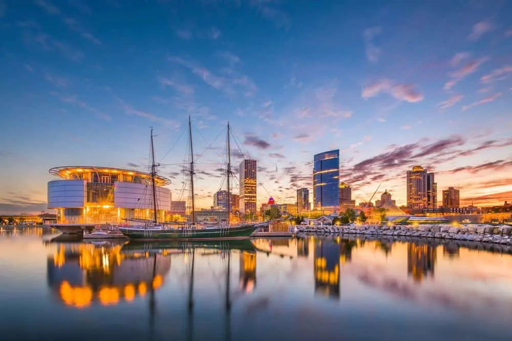 October might be the best time to visit milwaukee, lake side view of Luxury Resort near Milwaukee with many tall buildings lit up by electric lights and an old ship with masts and rigging moored in front all under a dramatic cloudy sky at dusk