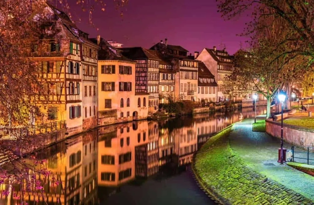 Find your favorite countries to visit near Luxembourg, view of the river in strasbourg by night with one side covered in densely packed residential buildings and the other with a footpath and trees lit by Victorian street lights