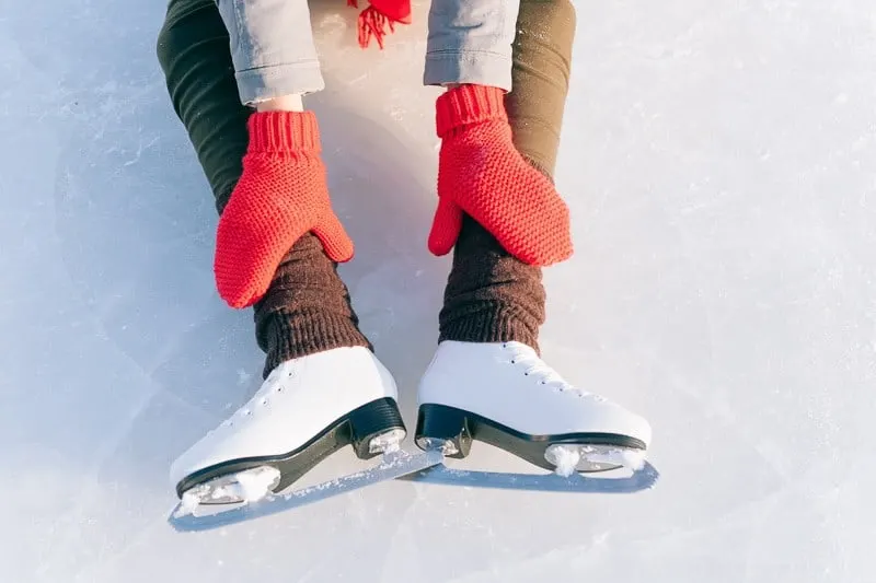 sitting person touching their ice skating boots, best things to do in lake geneva in december, young girl with mittens in ice skates sitting on ice and leaning forward