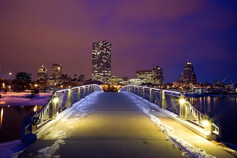 best things to do in Milwaukee in January, View of city skyline seen across a bridge covered in a light layer of snow and lit up with soft yellow lights under a purple and blue sky at night