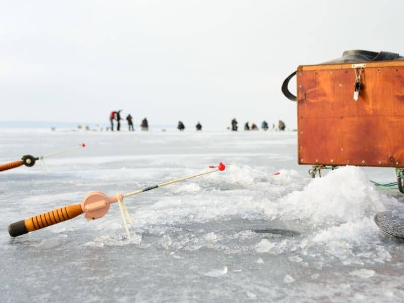 Sturgeon Bay winter events, Close up shot of ice fishing rods left above circular holes cut in a frozen lake next to a wooden box of ice fishing supplies with a line of people visible in the background on a grey cloudy day