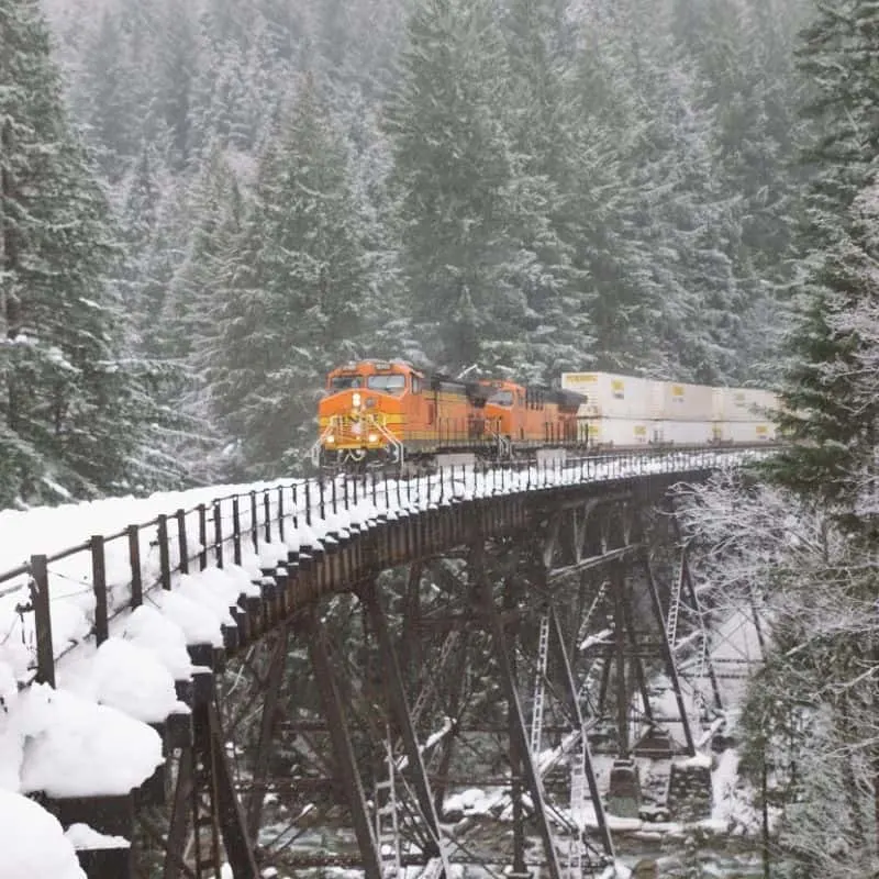 orange train driving over metal bridge in the snow surrounded by snow covered pine trees