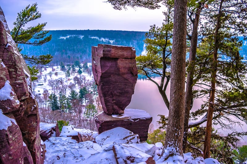 A large rock positioned on top of another large rock surrounded by tall thin trees on the side of a hill overlooking a lake with rolling hills covered in green trees stretching off into the distance all covered in a layer of soft white snow under a cloudy grey sky