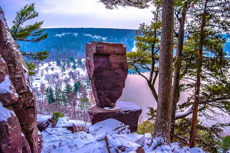 winter campgrounds in Wisconsin, A large rock positioned on top of another large rock surrounded by tall thin trees on the side of a hill overlooking a lake with rolling hills covered in green trees stretching off into the distance all covered in a layer of soft white snow under a cloudy grey sky