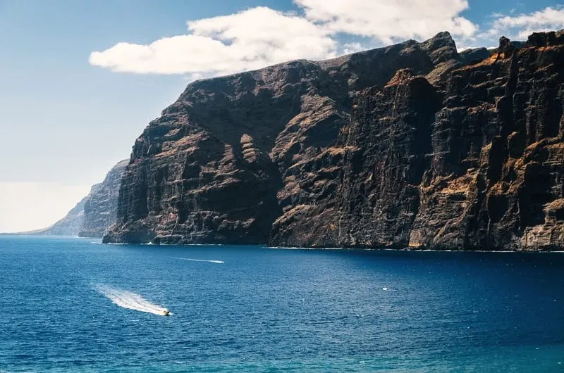 view of large rocky cliffs standing on the coast next to wide open blue ocean with the distant figure of someone riding a jet ski in the foreground