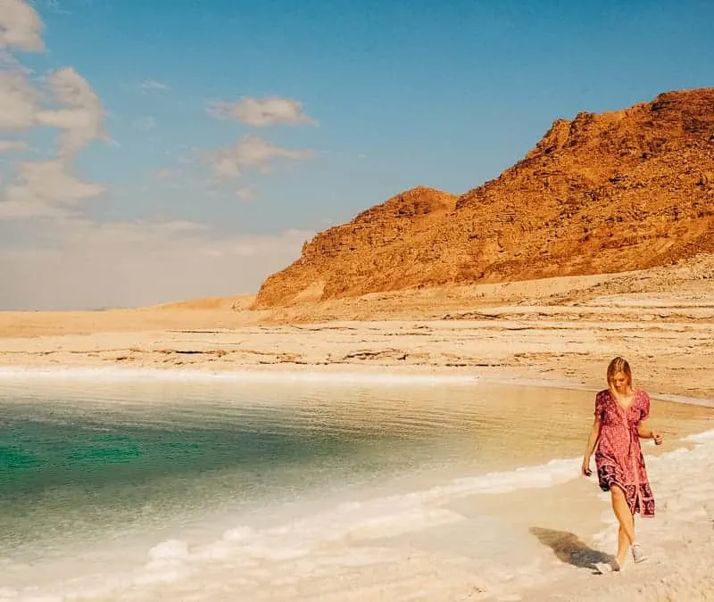 things to do in jordan, hiking to wadi mujib near dead sea, jordan, a woman walking by the beach with a mountain on the background