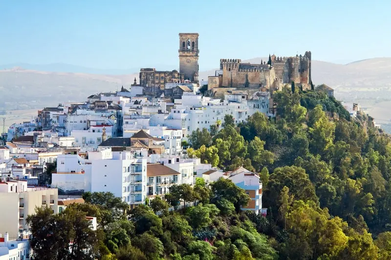 Discover the wonders of the paradores of spain, view of the arcos de la frontera with large stone castle and tower sitting above a built up area of white residential buildings on top of a hill covered in thick green trees with rolling hills behind