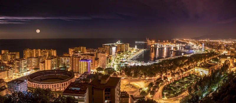 Stay in some of the oldest hotels in spain, view of malaga at night with tall residential buildings mixed with older structures and tree-lined roads next to the ocean with a full moon in the sky