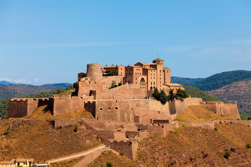 Select your next destination from a map of paradores in spain, view of large fortification with many towers and turrets sitting inside a series of stone walls atop a hill with rolling hills behind