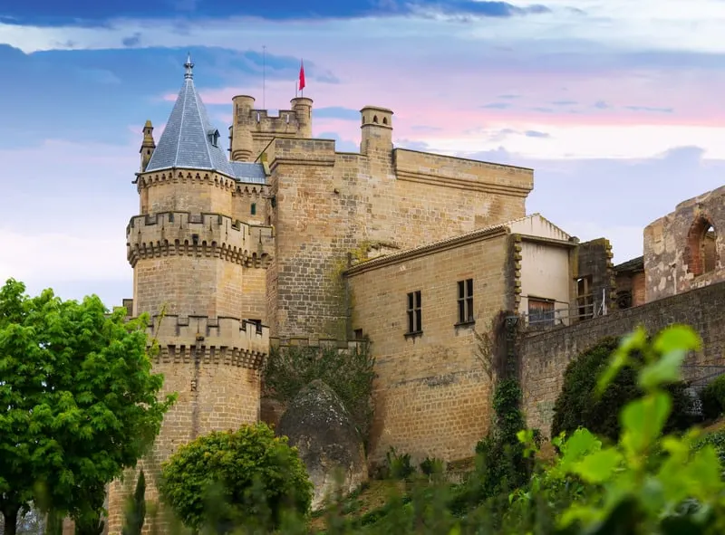 Stay in some of the most opulent government hotels in spain, view of the Castle of Olite with tall stone walls and pointed turrets under a pink sky at sunset