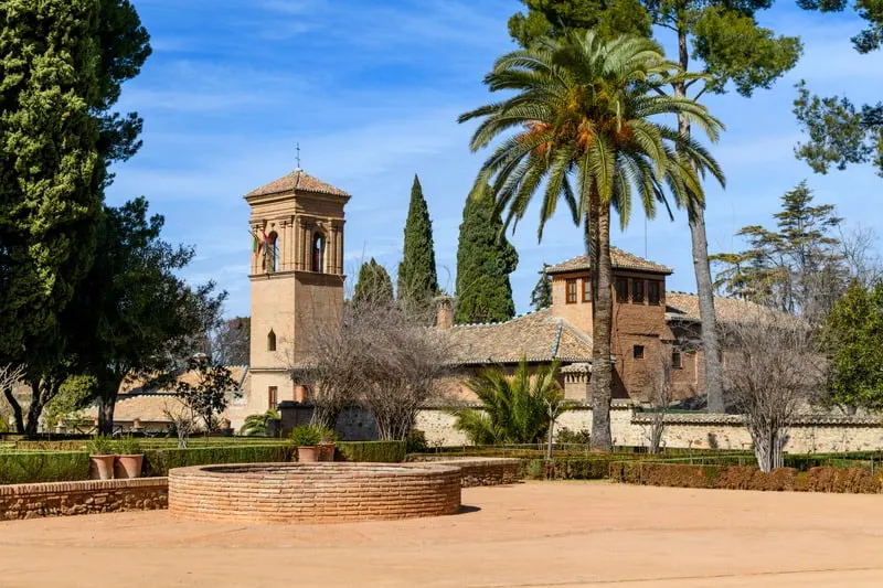 Stay in these parador hotels spain this year, courtyard with low walls and hedges and small circular well with tall green trees and the Convento de San Francisco parador building behind with its stone towers and tiled rooftops under a blue sky