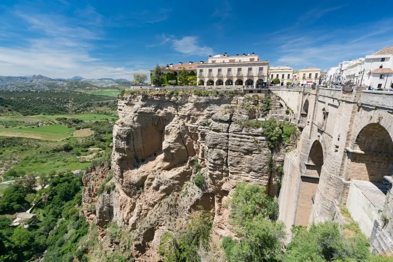 Enjoy the best paradores spain has to offer, view of Parador de Ronda sitting on top of rocky cliff next to large stone bridge with rolling green fields and trees below under a wide blue sky