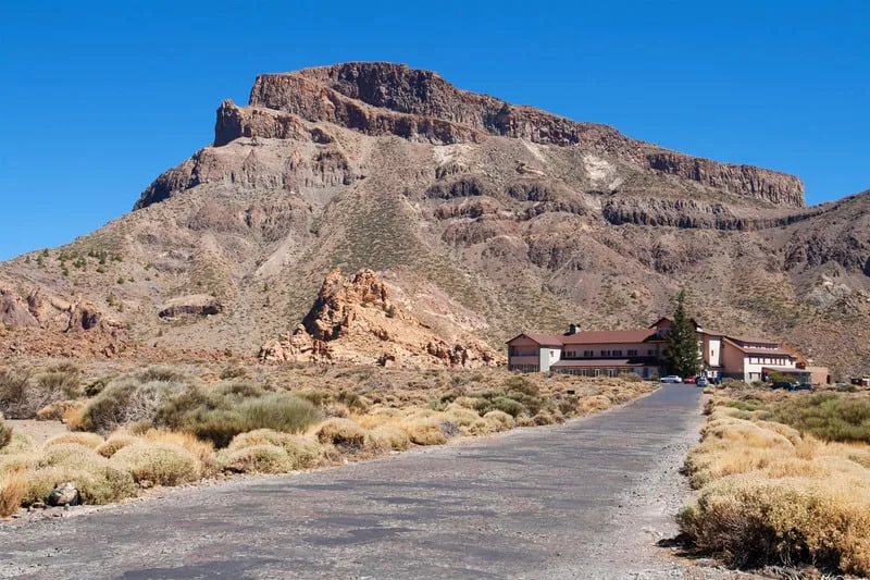 Discover the best parador hotels northern spain can provide, long road leading to modern hotel structure sitting beneath a tall and imposing rocky mountain under a clear azure blue sky