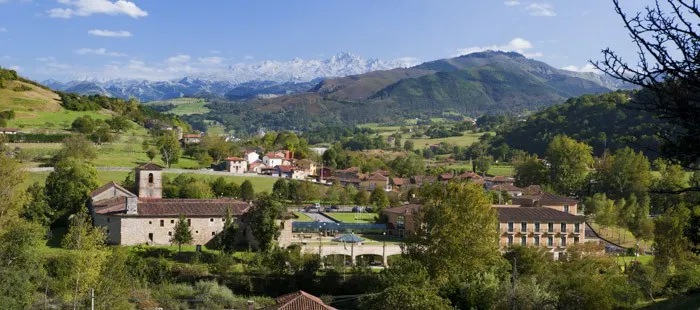 These spanish government hotels are some of the best in the world, view of idyllic Spanish countryside with older farm buildings and cottages surrounded by rolling green fields and clusters of trees with mountain range behind all under a blue sky with white clouds