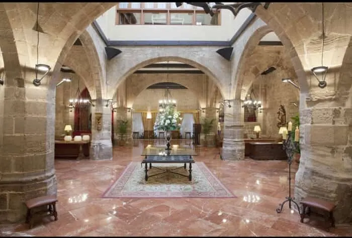Stay at some of the best spanish hotels in historic buildings, view of hotel interior with tall vaulted stone ceiling held up by stone pillars with a smooth tiled floor with ornaments and fine furnishings all around