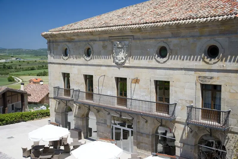 Check off all your favorites on this list of paradores in spain, view of Argomaniz Parador with large stone walls under a tiled rooftop with balcony doors and terrace with tables and chairs below with rolling green fields beyond