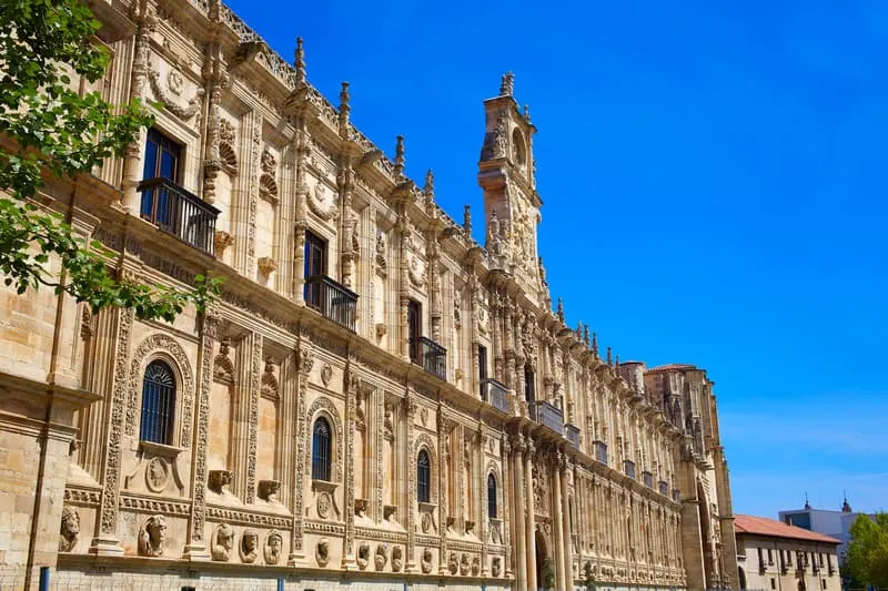 Don't miss out on any of these amazing spanish paradors, view of the Parador San Marcos in Leon with large stone walls covered in ornate carvings as well as a tall stone bell tower under a clear blue sky