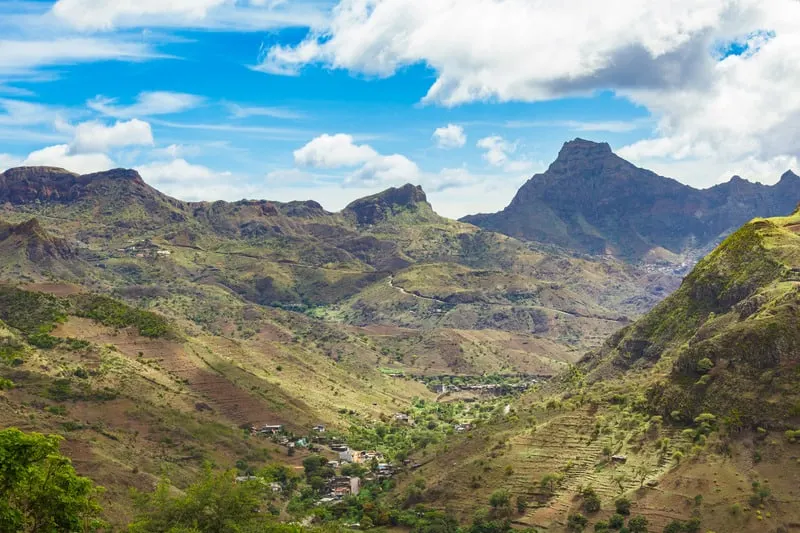 Try out all the things to do in cape verde boa vista, view of majestic valley with small collection of buildings dwarfed by rolling grassy hillsides and rocky mountain peaks all under a dramatic blue sky with white clouds