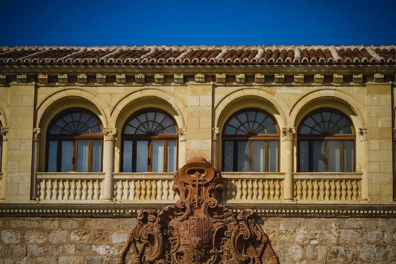 Enjoy the spa feeling of the paradores of spain this summer, close up view of the exterior of an ornate stone building with intricate carving underneath a set of arched windows beneath a tiled rooftop