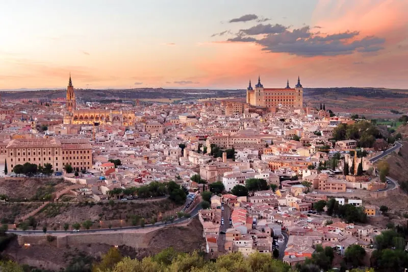 Try out the best paradores in Spain this year, view of city with tall older structures with towers sitting in a sea of densely packed houses and roads under a bright pink sky at sunset