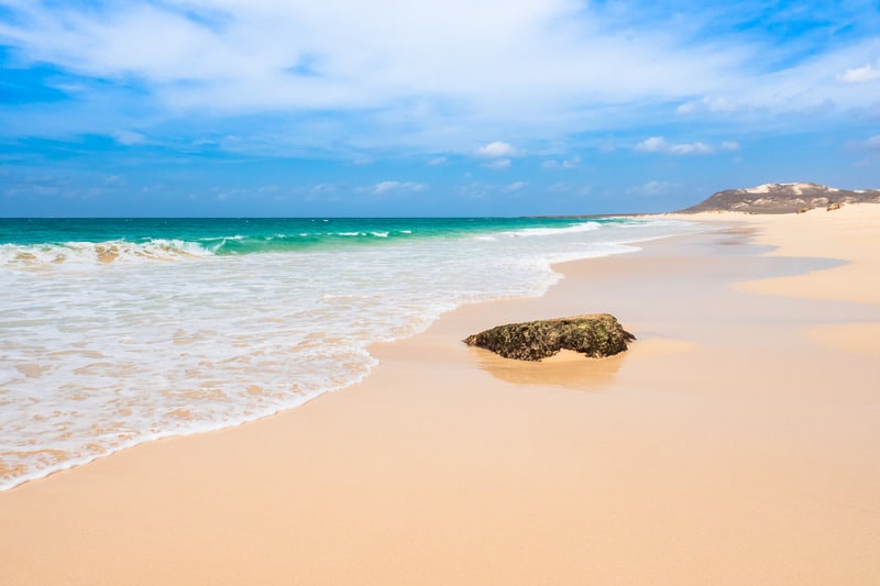 lone rock sitting on shores of deserted sandy beach next to the white surf of a wave retreating into the vibrant turquoise waters of the ocean under a bright blue sky with wispy white clouds