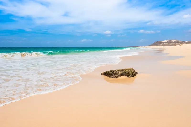 Enjoy some shopping in boa vista, cape verde, lone rock sitting on shores of deserted sandy beach next to the white surf of a wave retreating into the vibrant turquoise waters of the ocean under a bright blue sky with wispy white clouds