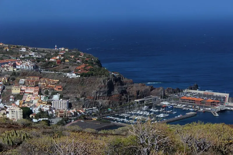 Try out a spanish parador on your next vacation, view of a coastal town with a harbor full of sailing boats and a series of buildings resting on a tall rocky hill next to the wide blue ocean