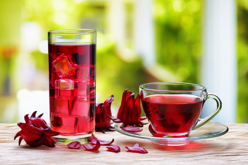 Cup of hot hibiscus tea (karkade, red sorrel, Agua de flor de Jamaica) and the same cold drink with ice cubes in glass on wooden table. Drink made from magenta calyces (sepals) of roselle flowers.