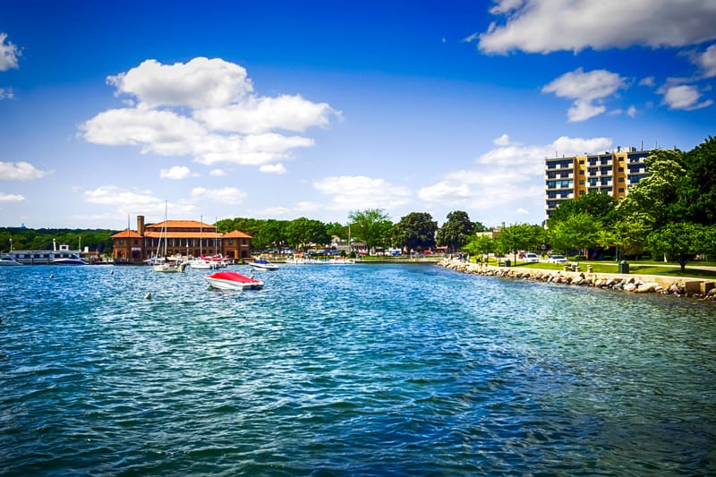 Fun things to do in milwaukee in fall for couples, view of large lake with small boats moored near to a lakeside building with green trees and park area to one side all under a bright blue sky with white fluffy clouds