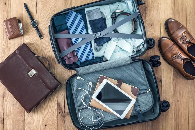 Open suitcase packed with clothing on one side and electronics in the other, beside suicase is a briefcase, wallet, watch and dress shoes