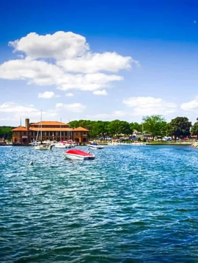 Wisconsin getaways in March, View from the water of a harbor with some small sailing boats moored next to a large orange building with some green trees behind all under a bright sky with clouds