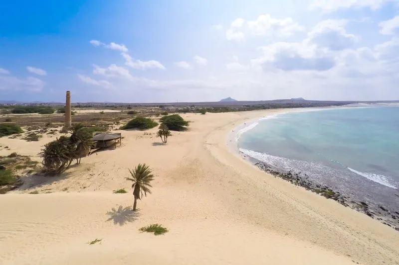 a beach with white sand and blue sea with a hut and a few trees on a bright day with clouds in the sky