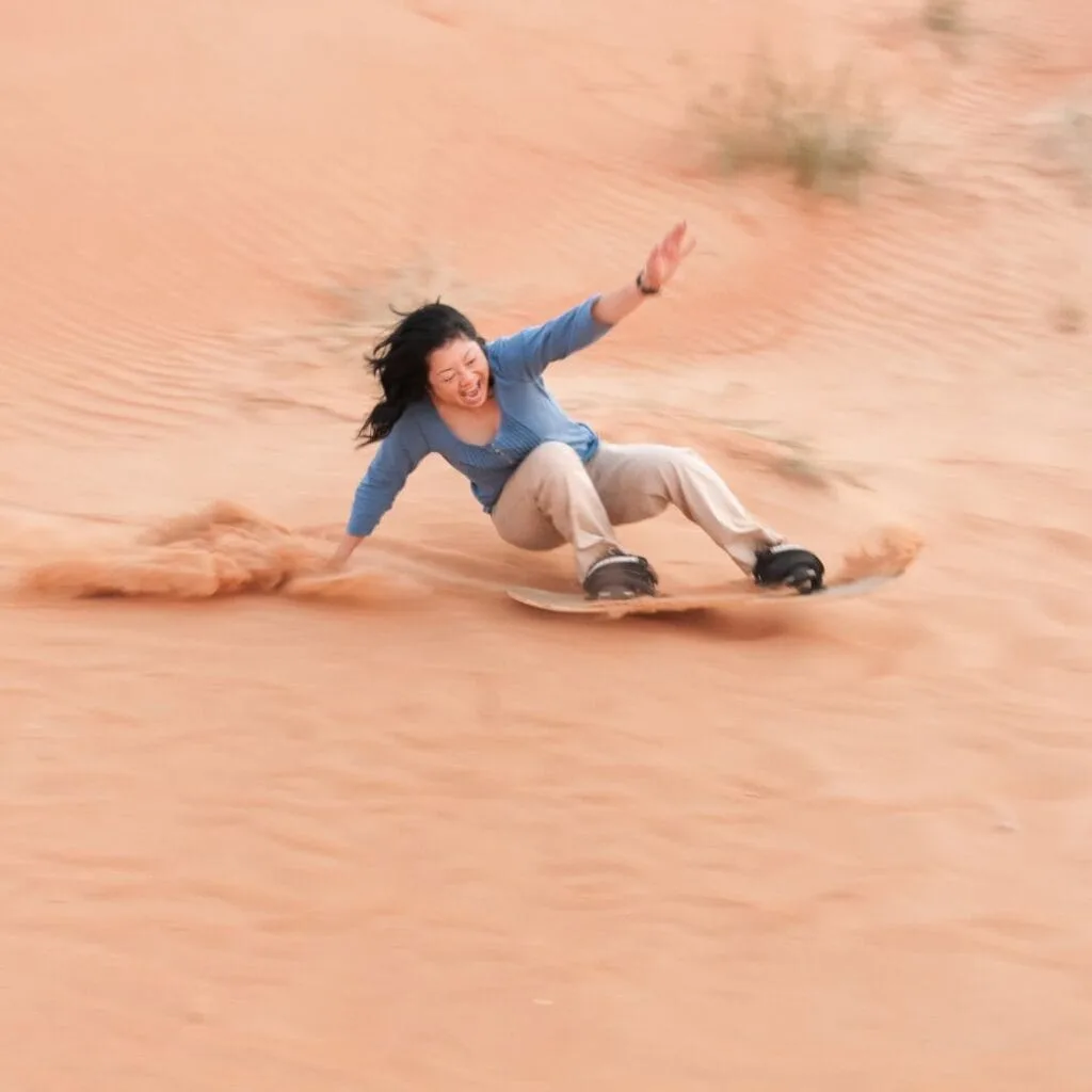 A happy, mature, Asian woman loses balance and is about to crash on her sandboard at the bottom of a sand dune in a desert