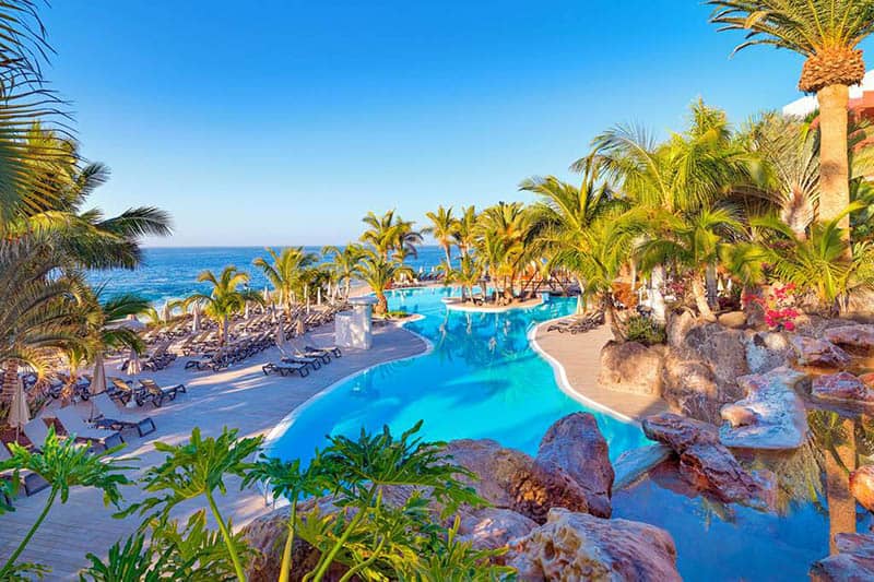 Enjoy these Tenerife resorts for families, Inside view of Adrián Hoteles with many green palm trees surrounding a large outdoor pool area with rows of sun loungers off to one side all under an empty blue sky