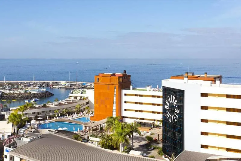 Head to the best resorts in Tenerife for families this summer, Top View of hotel BeLive Experience La Nina with some green palm trees and swimming pools visible below and a large view of the sea in the distance