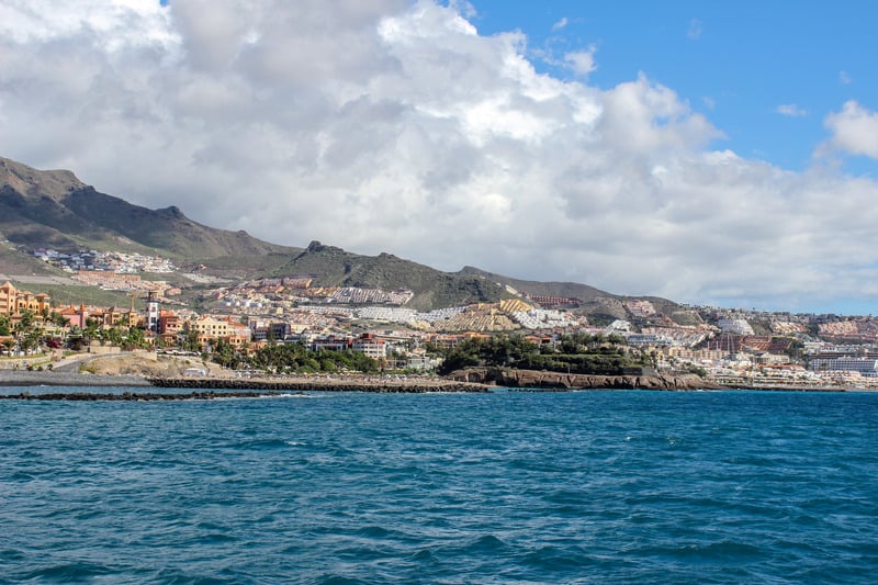 Stay at possibly the best resort in Tenerife for young families, View on the beach of Costa Adeje, Tenerife with rows of houses and other buildings leading up the slow slope of the hill underneath a bank of heavy white clouds