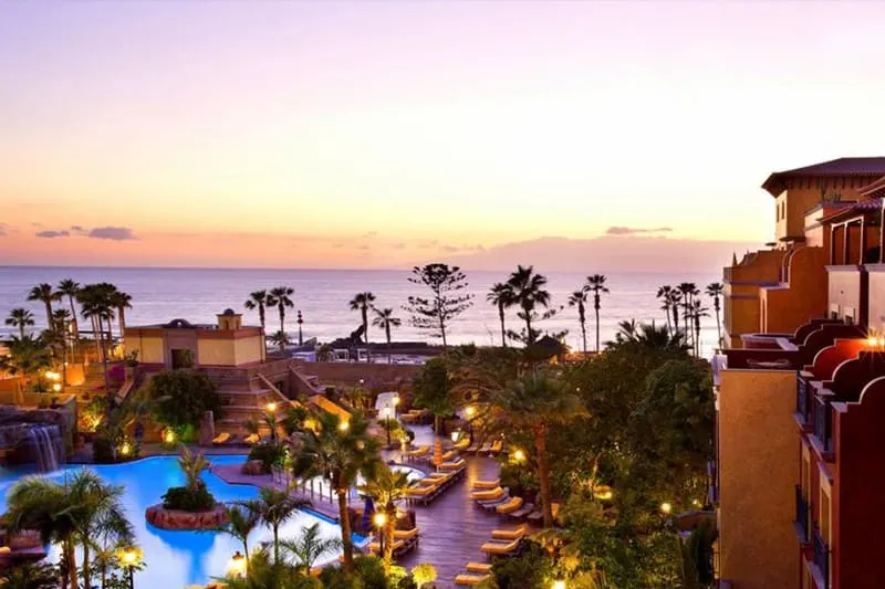 Discover the best place in Tenerife for families, A view of the pool and hotel at Europe Villa Cortes GL with areas of dense greenery below and the flat calm sea visible in the distance leading towards a bank of soft grey clouds at sunset