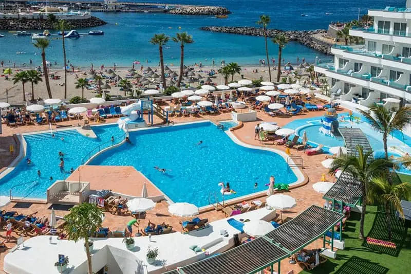 Enjoy some of the best places in Tenerife for families, A view of the pool at HOVIMA La Pinta Beachfront Family Hotel or Beach view with many beach umbrellas covering people laying on sun loungers with the beach and a small harbour visible behind