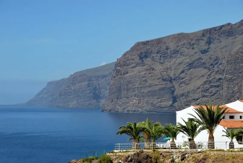 Head to the best Family Hotels in Tenerife this year, View of  los gigantes canyons, Spain with tall rocky cliffs sitting by the blue waters of the ocean with some houses and palm trees in the foreground