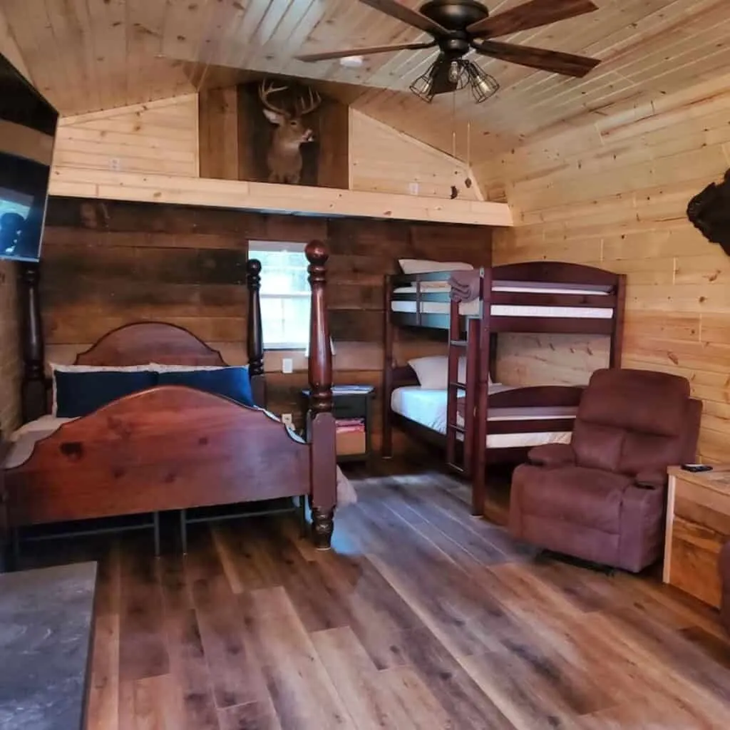 inside a wooden cabin with a big queen bed and a double deck bed with a hanging deer on the wall