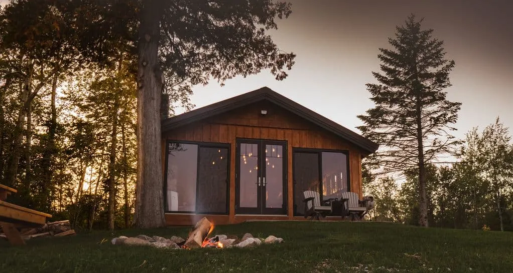 a view of a cabin in the woods during sunset while there is a fire pit outside