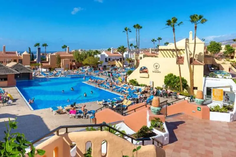 Don't miss out on these family friendly resorts in Tenerife, Inside hotel view  with outdoor pools and plenty of sunbathers lying on sun loungers with steps leading down into the pool area all under a wide open blue sky