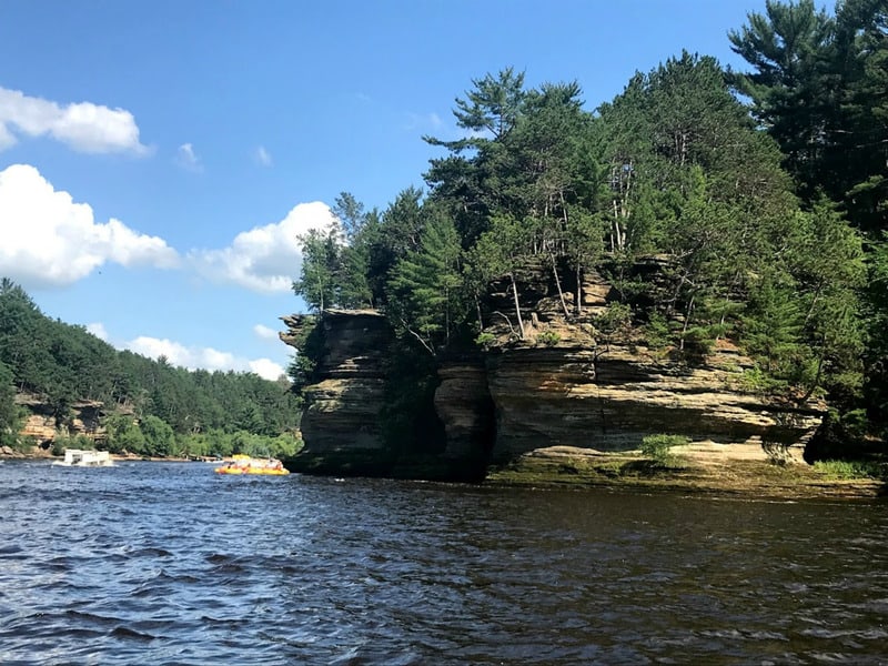 See the best destination for fall Wisconsin getaways, view of a rock formation covered in green trees and shrubs next to a body of water at the Wisconsin Dells all under a bright blue sky with some white clouds