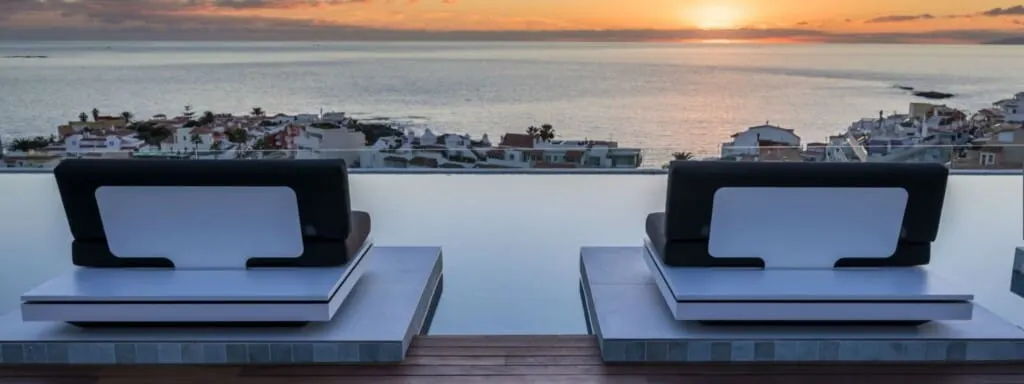 Check out these Romantic Hotels In Tenerife places, view from behind of two sun loungers facing out across a wide view of some buildings next to the sea at sunset