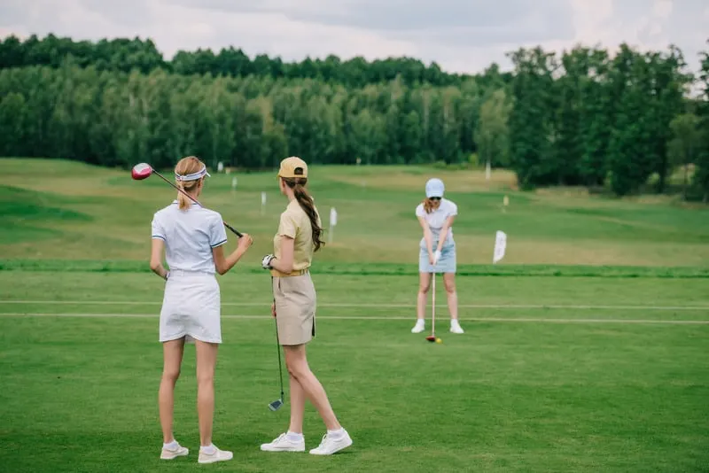 selective focus of women in caps with golf equipment looking at friend playing golf at golf course in Wisconsin