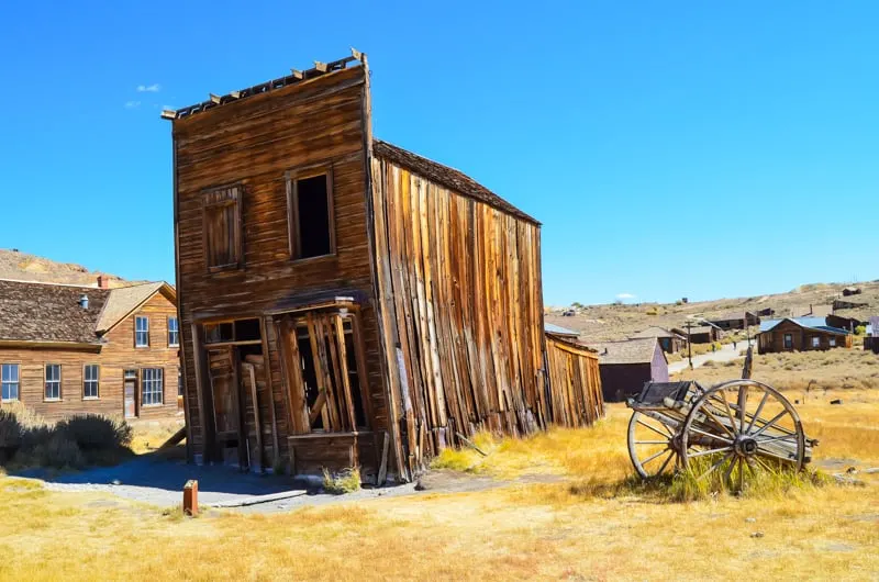 Check out abandoned places in united states, tall wooden building leaning to one side with boarded up windows and an abandoned wooden wagon next to it