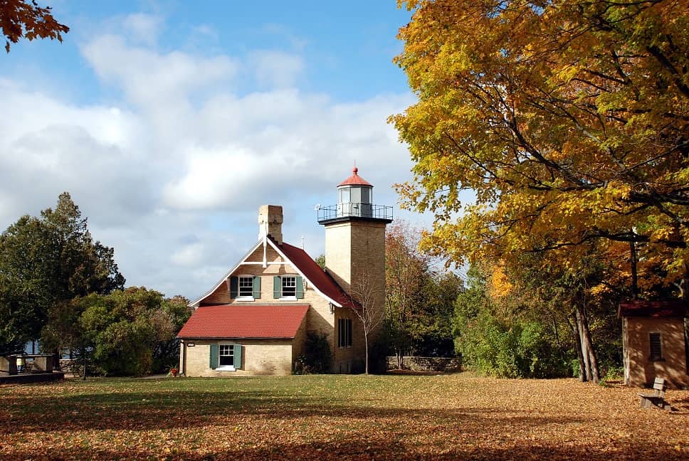 View of Eagle Bluff Lighthouse with single tower and small living quarters nearby with trees in fall colors to either side and a lawn of green grass partially covered in fallen leaves in front